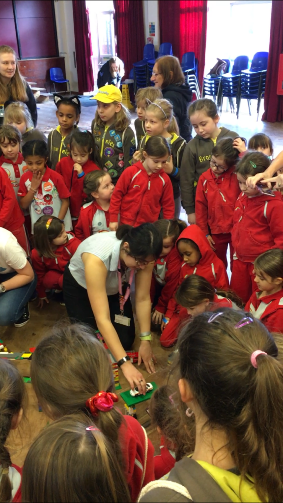 Brownies and Rainbows gathered round different model bridges made of Lego. A woman sits in the middle of the group testing the bridges with a toy car