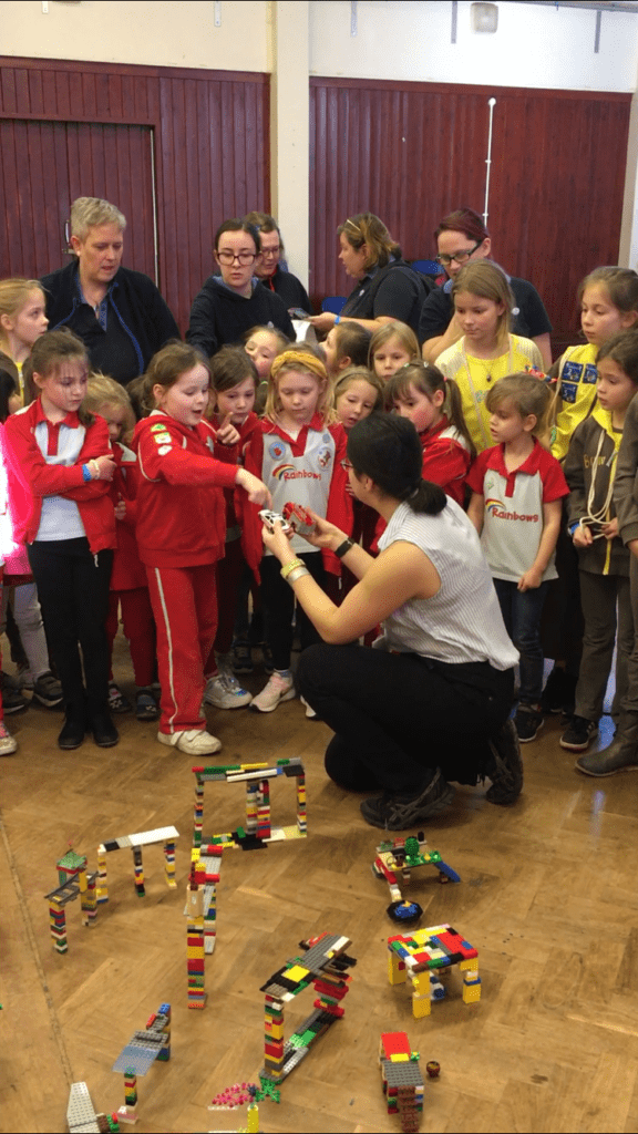 Brownies and Rainbows gathered round different model bridges made of Lego. A woman sits in the middle of the group and a Rainbow selects a toy car for the woman to test the bridge with